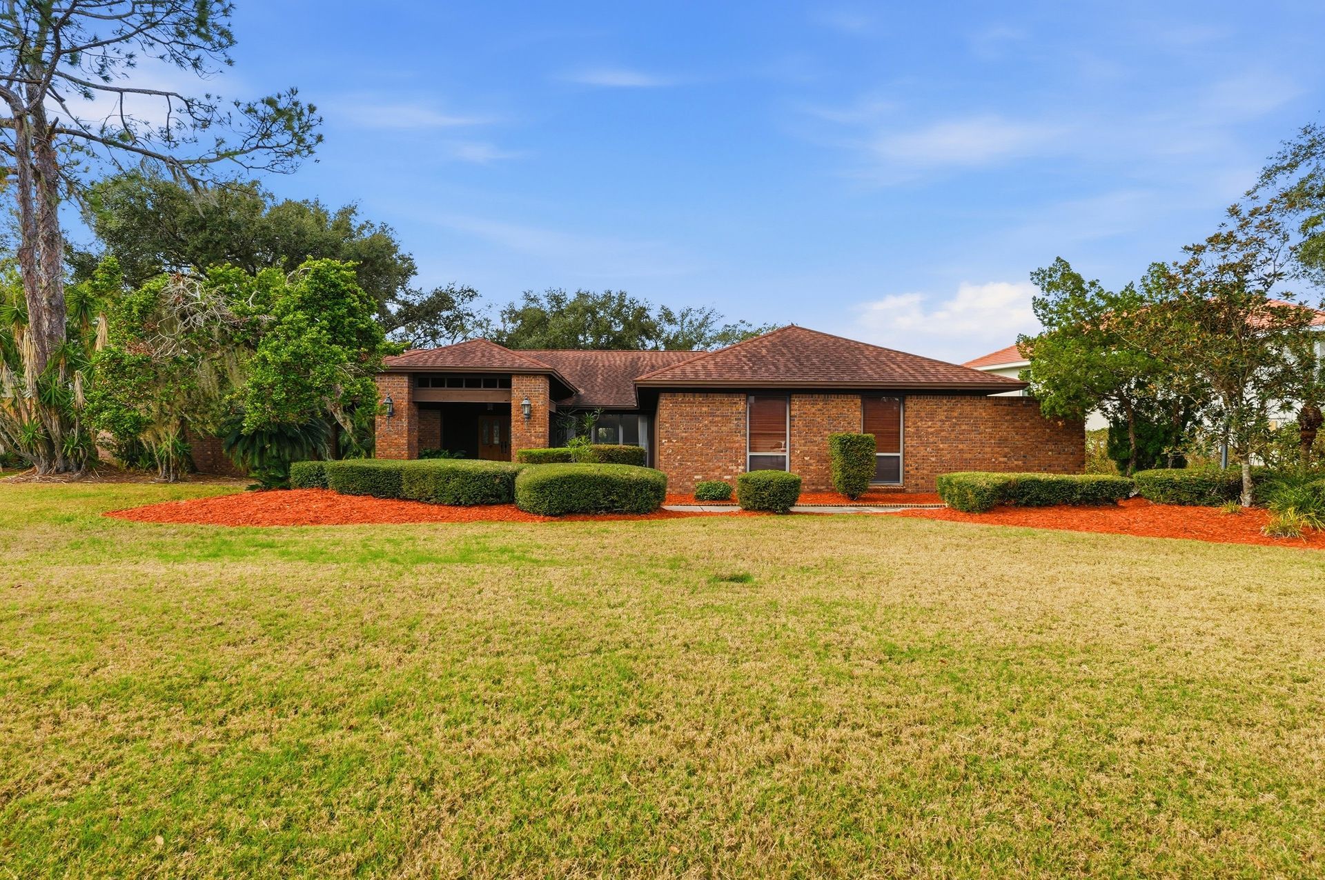 Single-story brick house with a red-tiled roof, front lawn, and flower beds under a blue sky