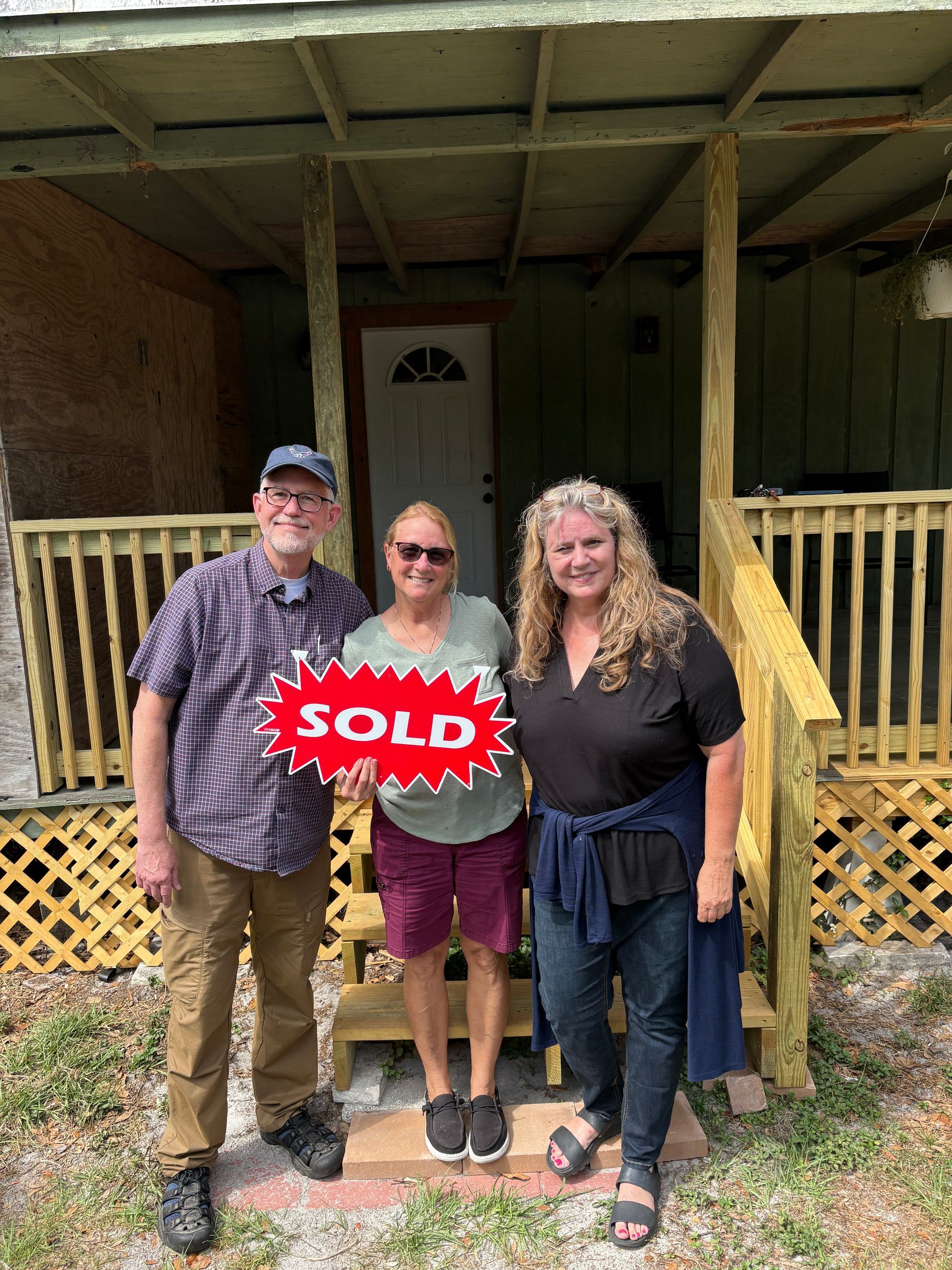 Three people standing on a porch holding a red “SOLD” sign
