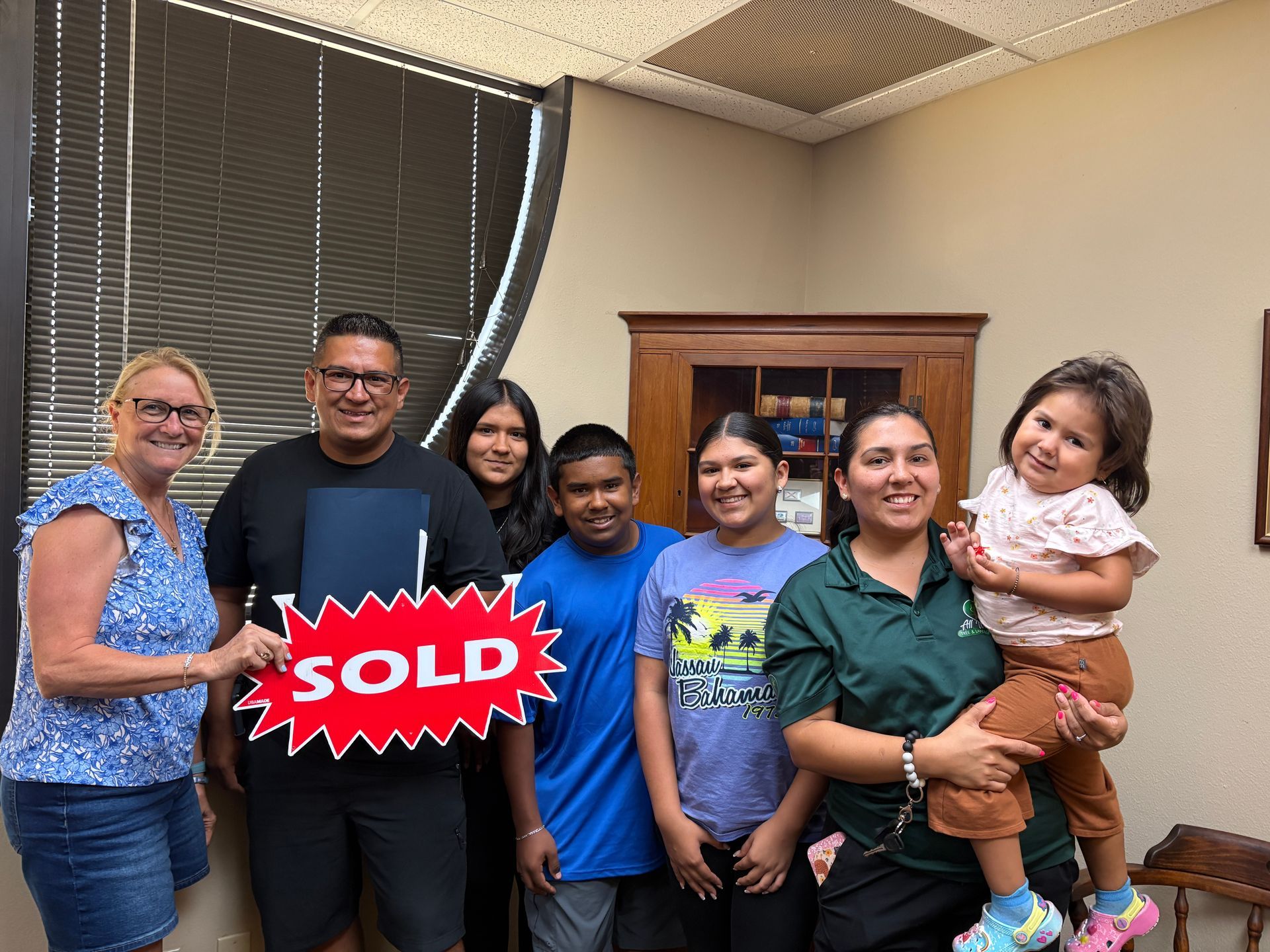 Group of six people smiling indoors, one holding a toddler, with a red “SOLD” sign in front