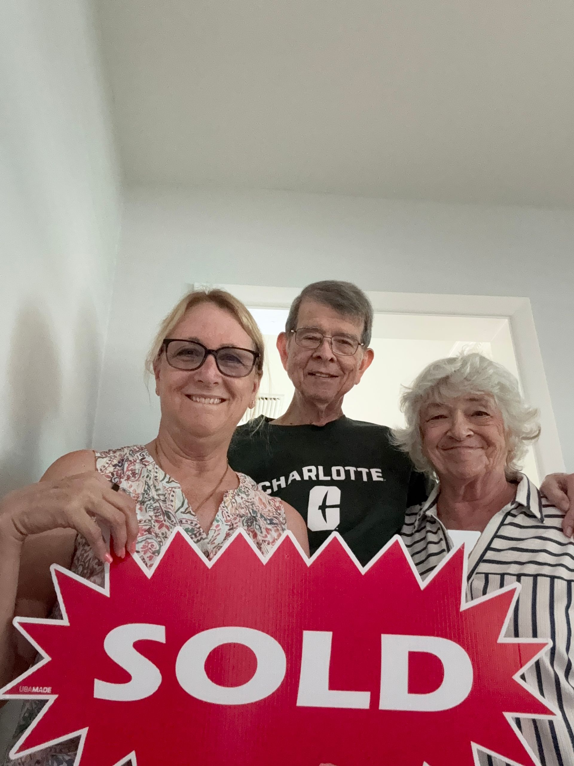 Three people holding a red “SOLD” sign indoors, smiling at the camera.