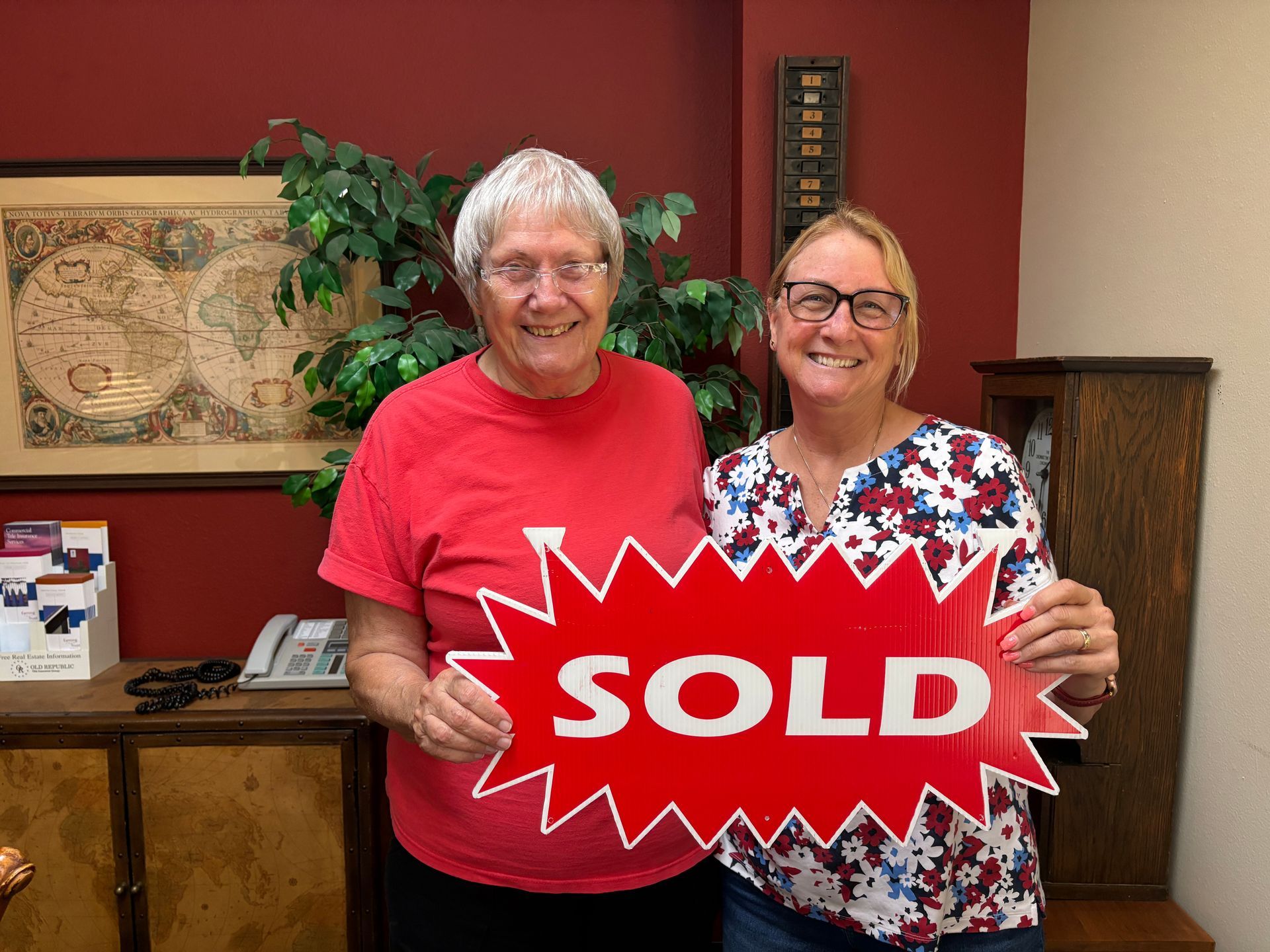 Two people smiling and holding a red SOLD sign in a room with a red wall and artwork.