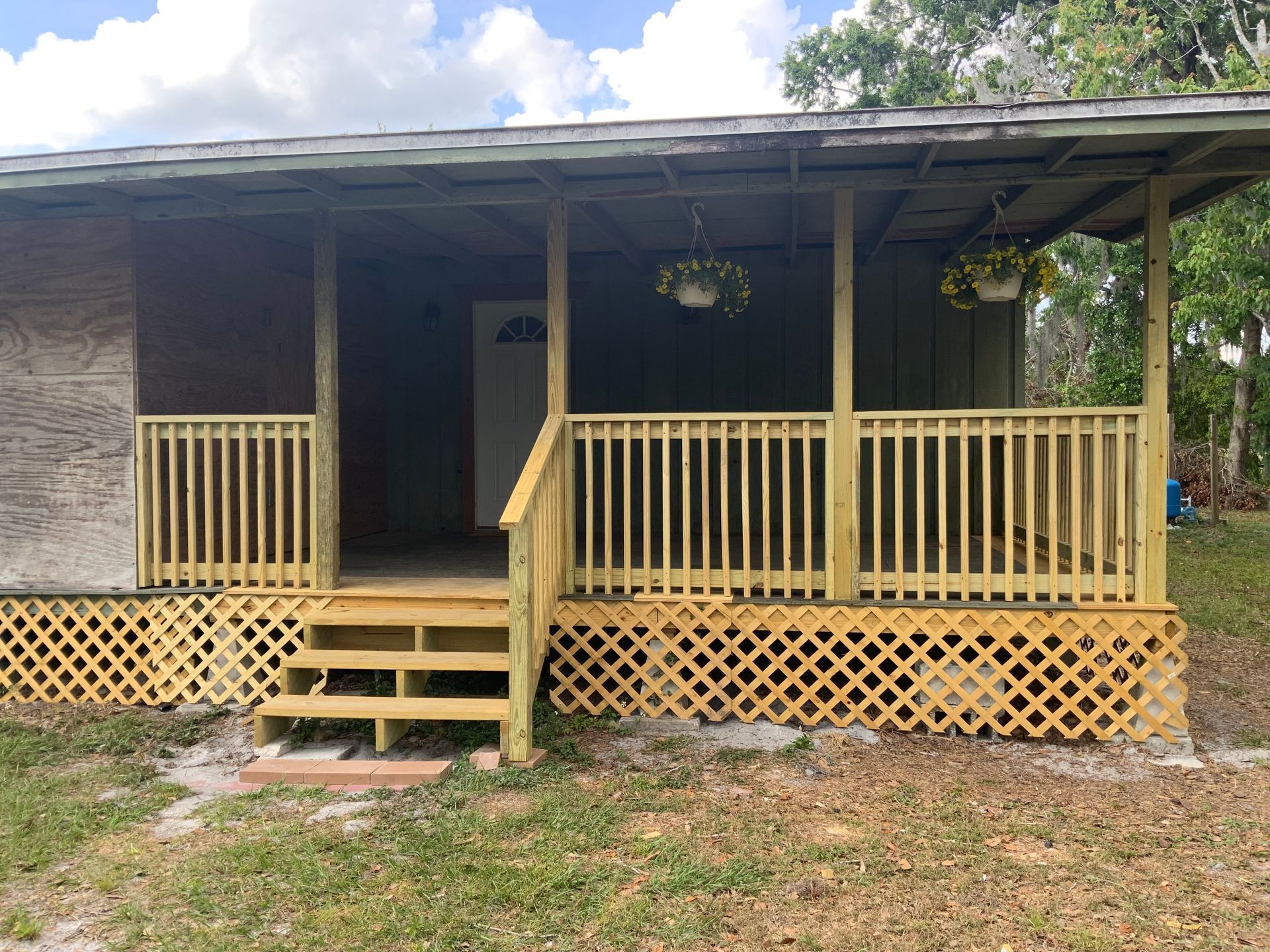 Small yellow porch on a rustic house with steps, railing, and lattice skirting under a cloudy sky