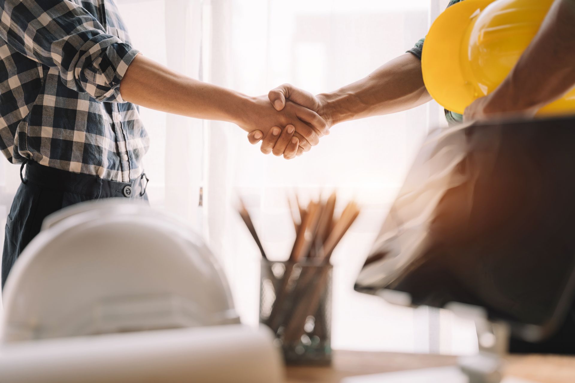 Two people shake hands across a desk in a bright office with a hard hat visible.