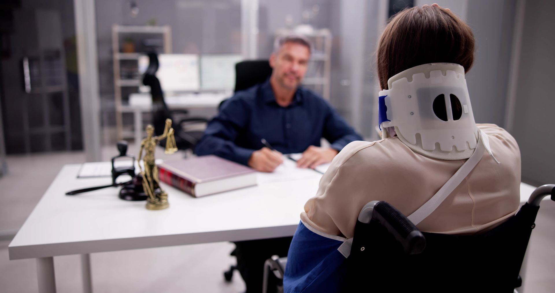 Two people in an office across a desk, one facing away in a white neck brace speaking to a man seated opposite.