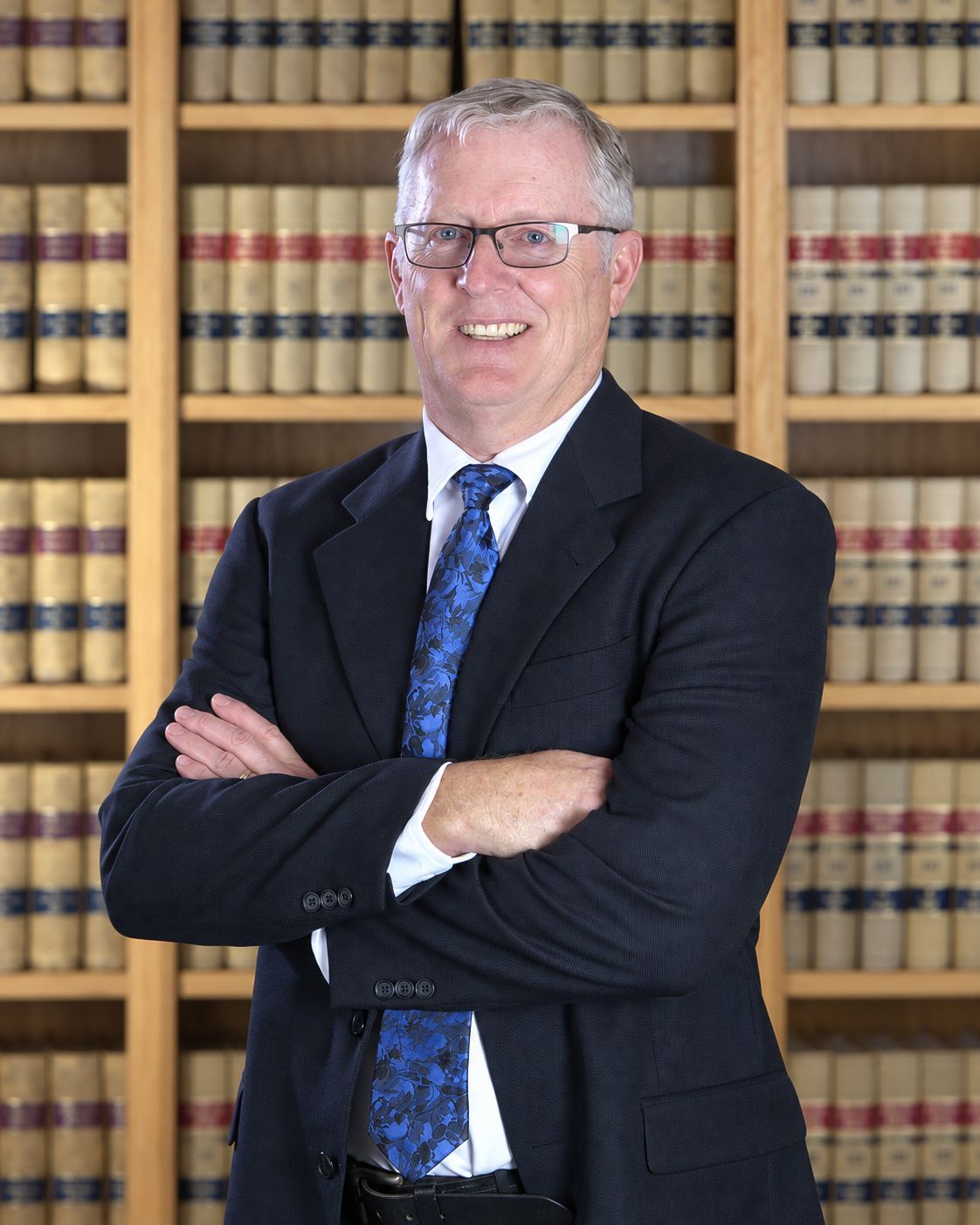 Smiling man in a black suit and light blue tie, standing with arms crossed against a blue studio backdrop