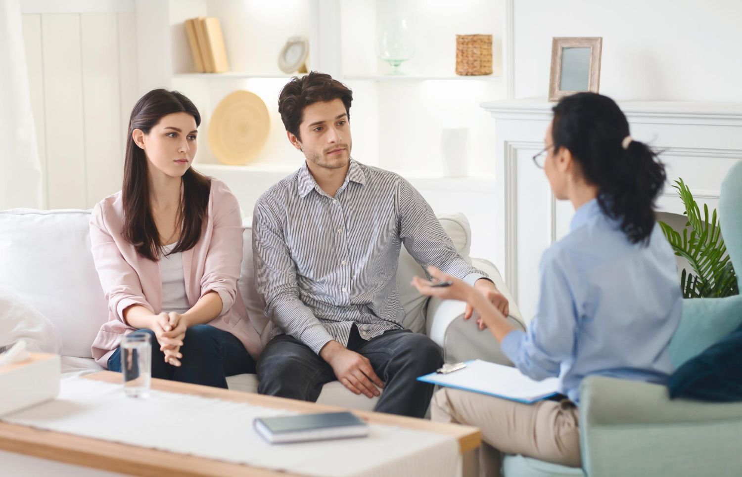 Three people in a counseling session on a couch, talking with a therapist in a bright living room.