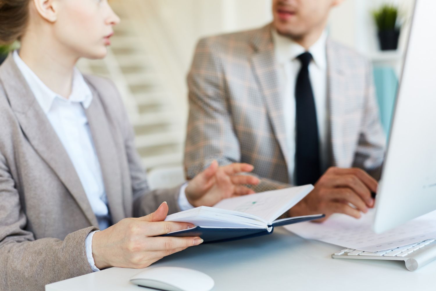 Two coworkers in light blazers discussing documents at a desk in a bright office