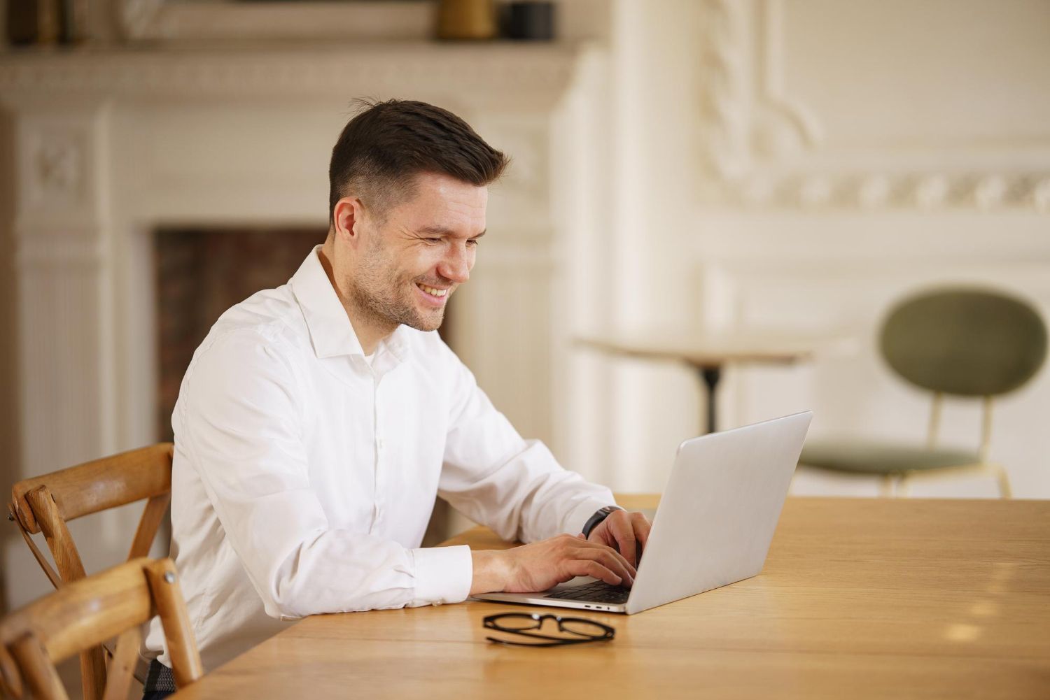 Man in a white shirt typing on a laptop at a wooden table in a bright room