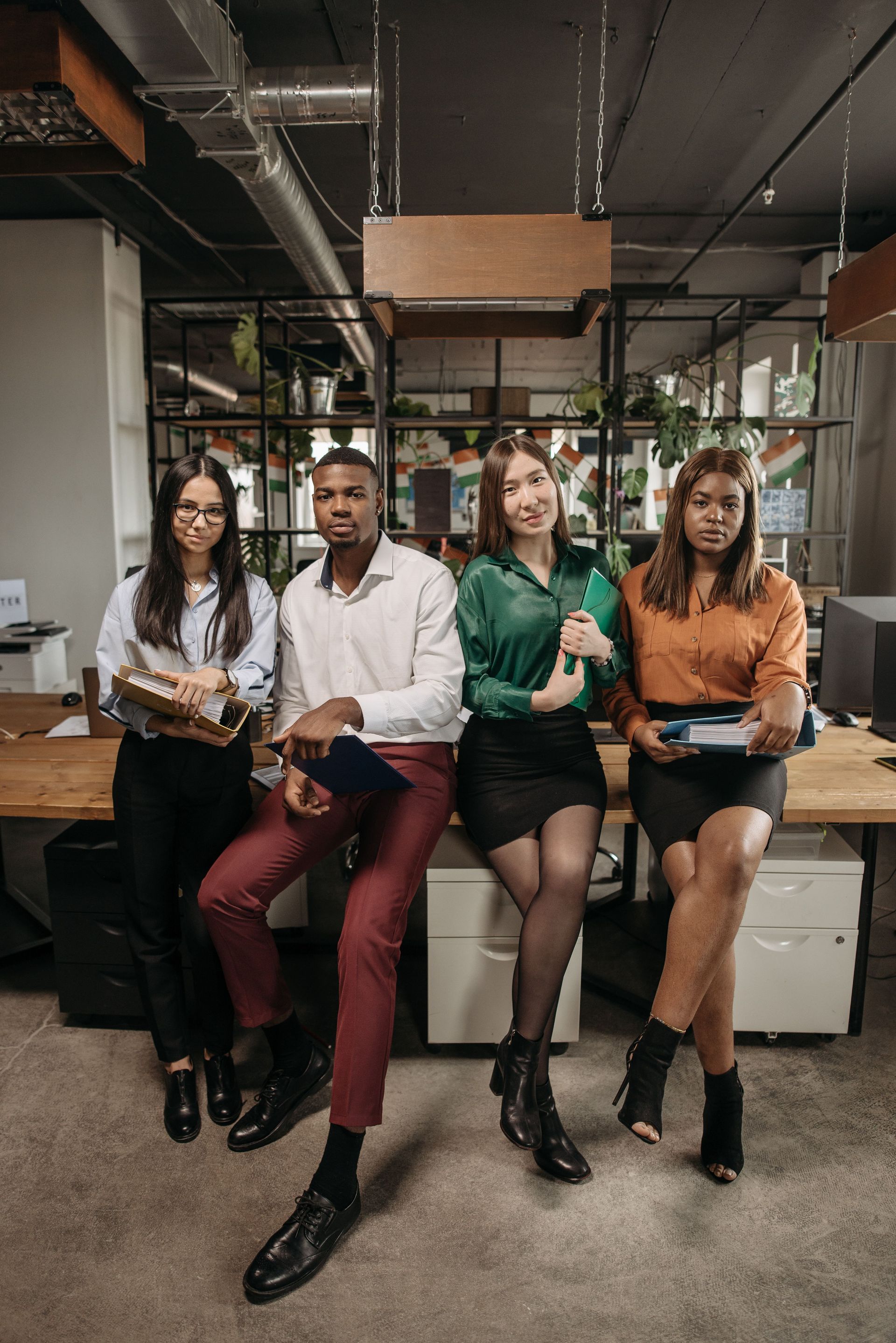 Four coworkers sitting on a workbench in an industrial office space, holding notebooks and a phone.