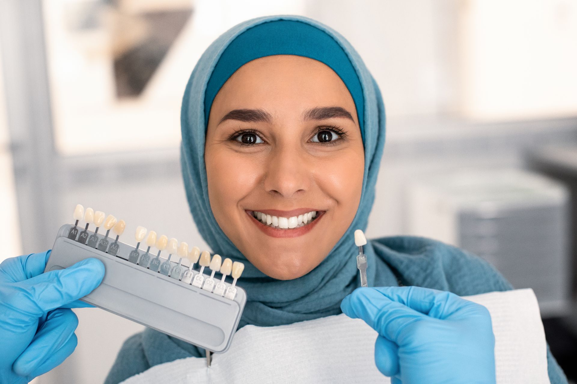 A woman in a hijab is smiling while holding a tooth color chart.