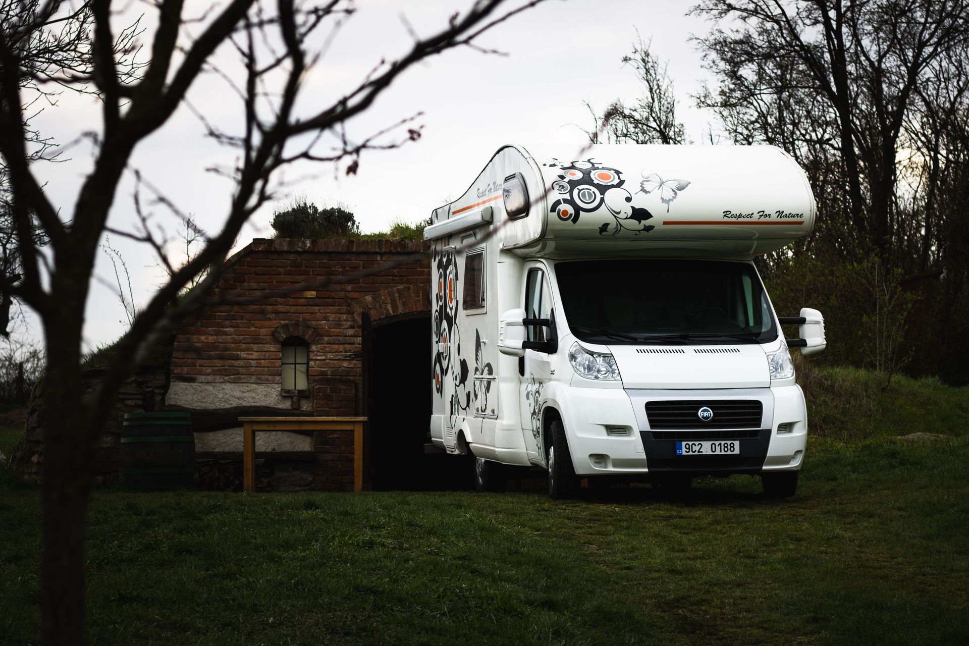 White camper van parked on grass in front of a small brick building with bare trees nearby