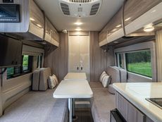 Modern RV interior with a long table, two benches, overhead cabinets, and side windows