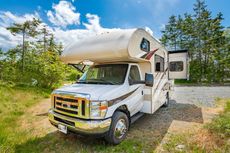 White camper truck parked on a gravel road beside green trees under a bright blue sky