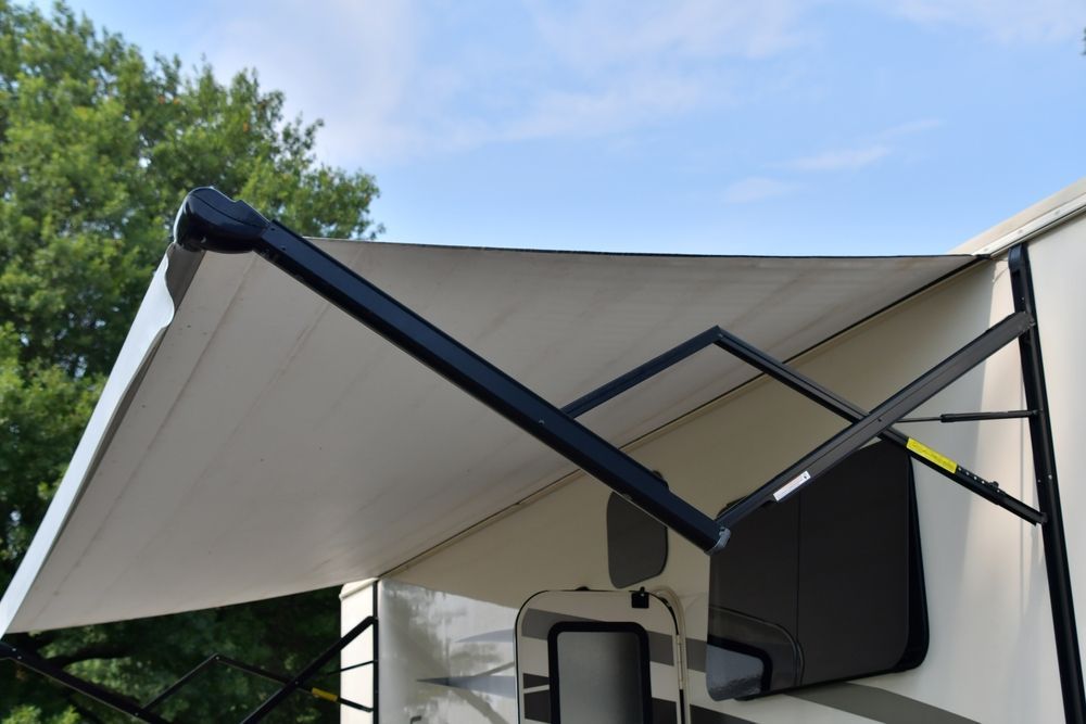 Retractable awning extended over a camper, with black support arms and a beige canopy under a blue sky