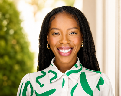 Portrait of a smiling person with long braids, wearing a white shirt with green abstract pattern, indoors.