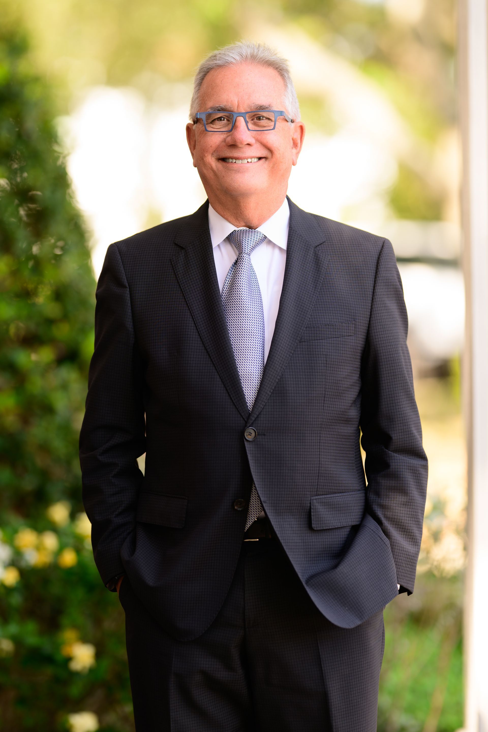 Smiling man in a dark suit and striped tie standing outdoors with hands in pockets, blurred greenery behind him