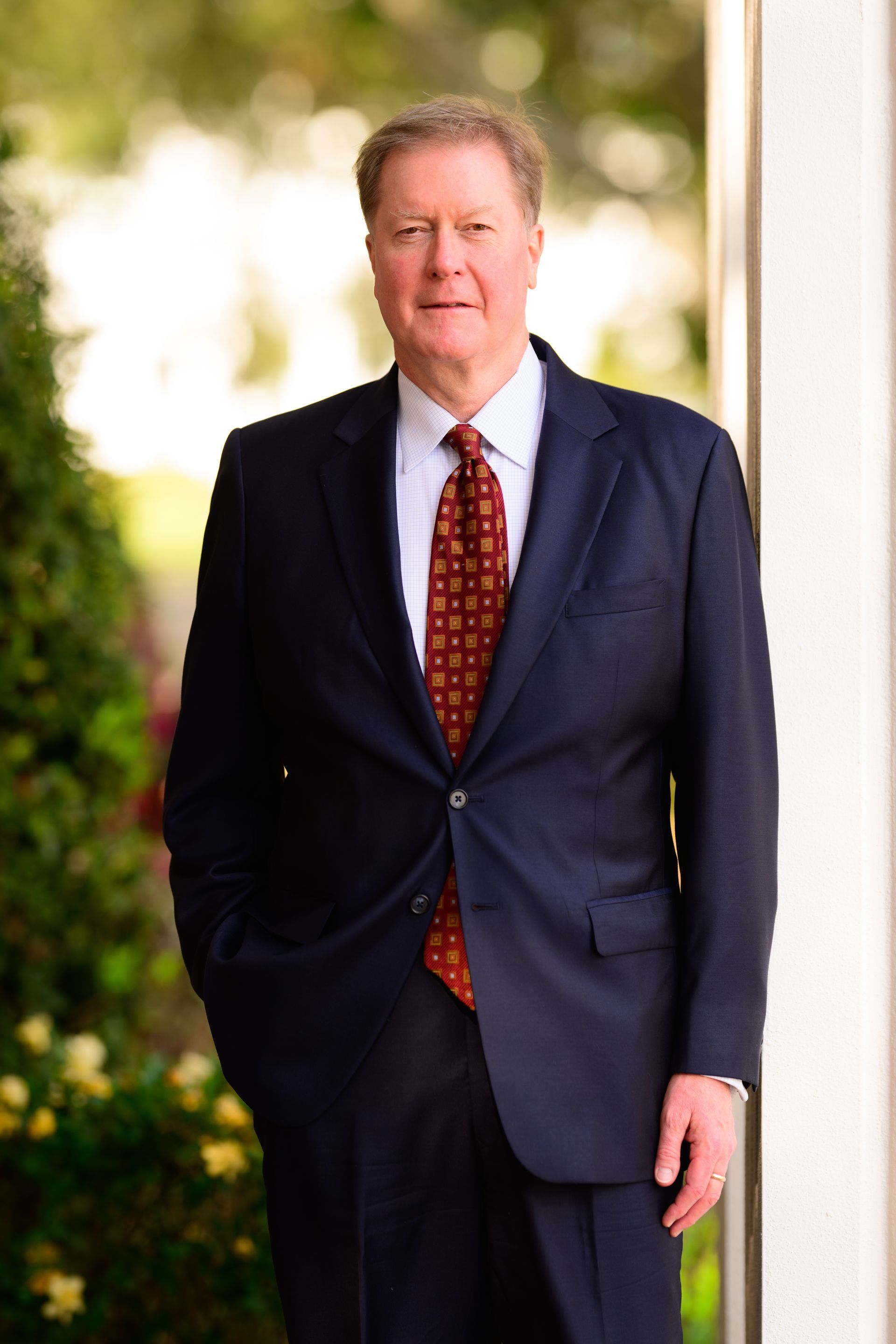 Man in a navy suit and red tie standing outdoors by a building column.