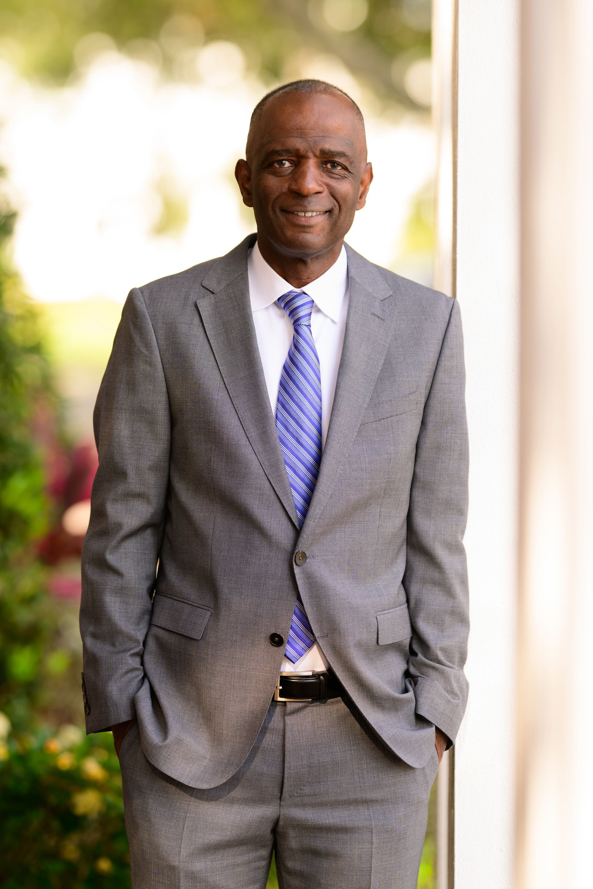 Man in a gray suit and blue tie standing outdoors with hands in pockets, smiling at the camera.