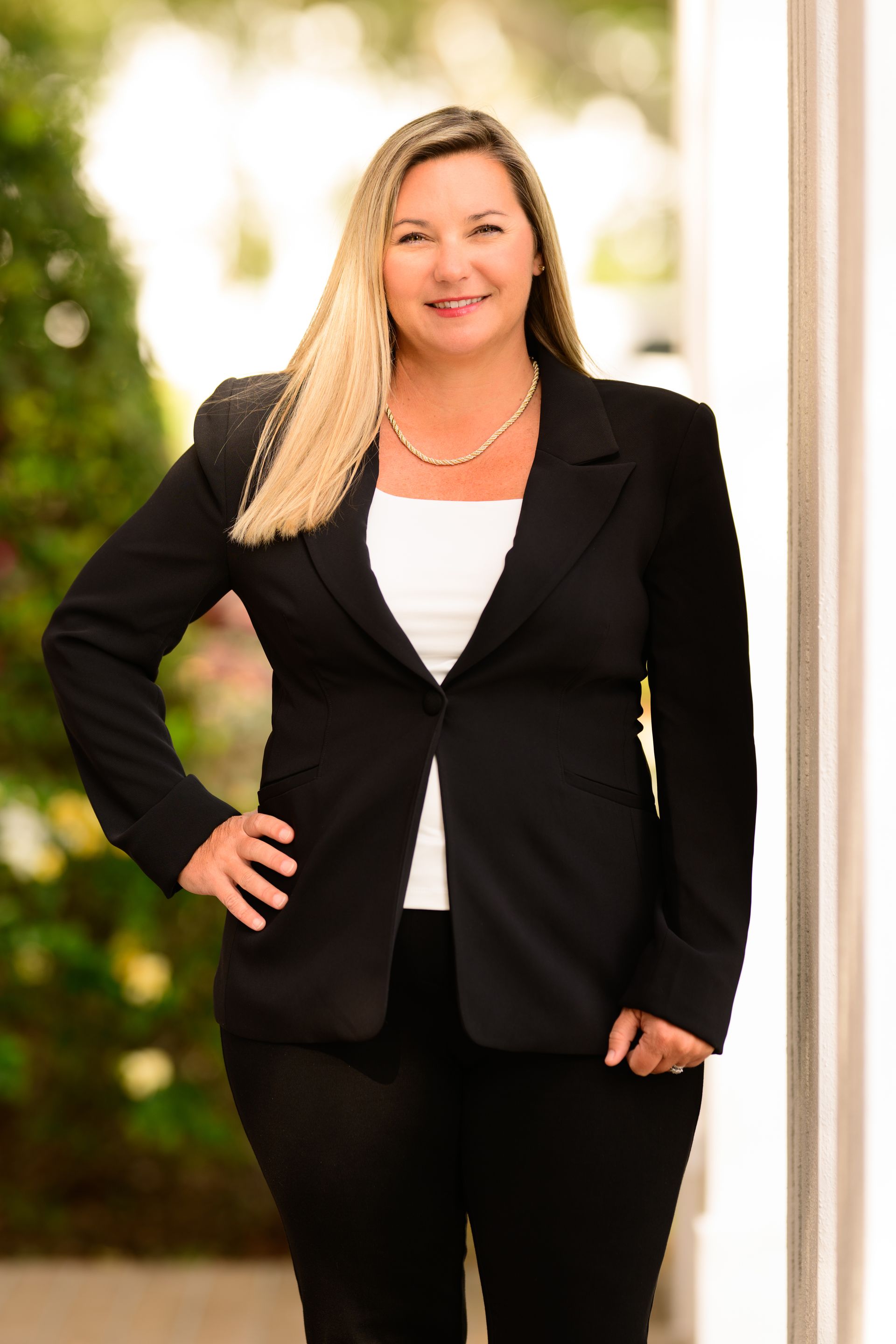 Woman in a black blazer standing outdoors with one hand on her hip, smiling at the camera.