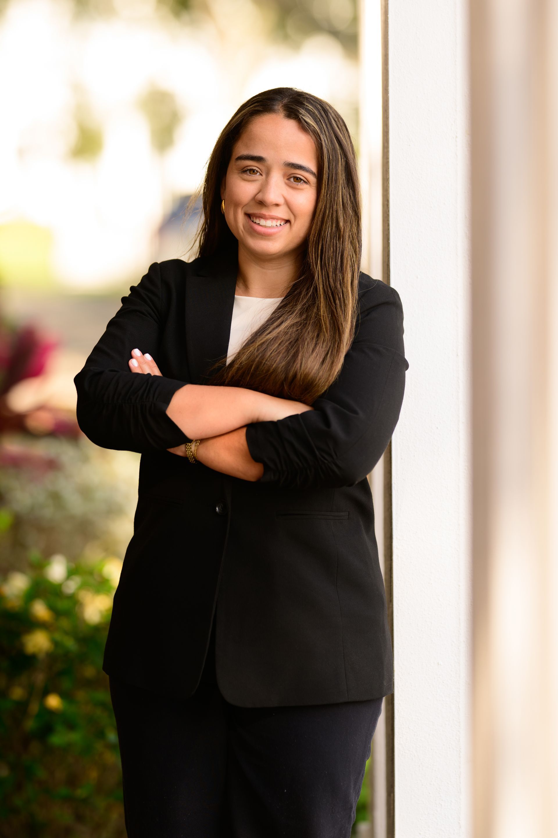 Woman in a black blazer stands outdoors with arms crossed, smiling near a column.