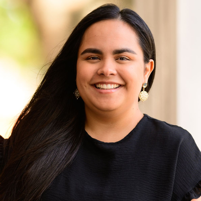 Smiling person with long dark hair in a black top against a softly blurred outdoor background