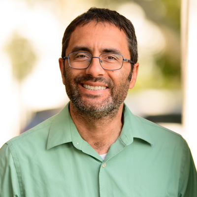 Smiling man in glasses and a light green shirt against a blurred outdoor background