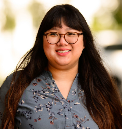 Smiling person with long dark hair and glasses in a gray patterned blouse, standing outdoors.