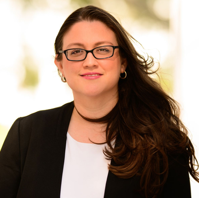 Portrait of a woman with glasses in a black blazer and white top, smiling against a bright background