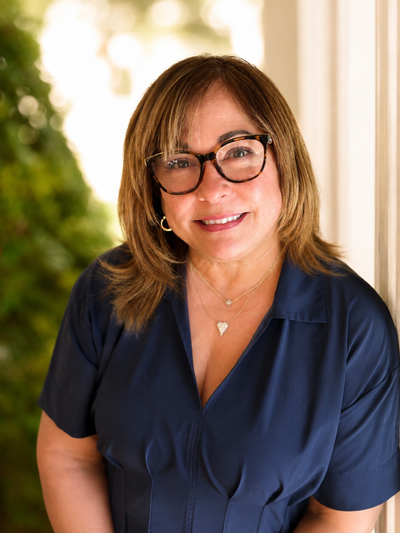 Smiling person in glasses and navy blouse standing indoors near a bright window.