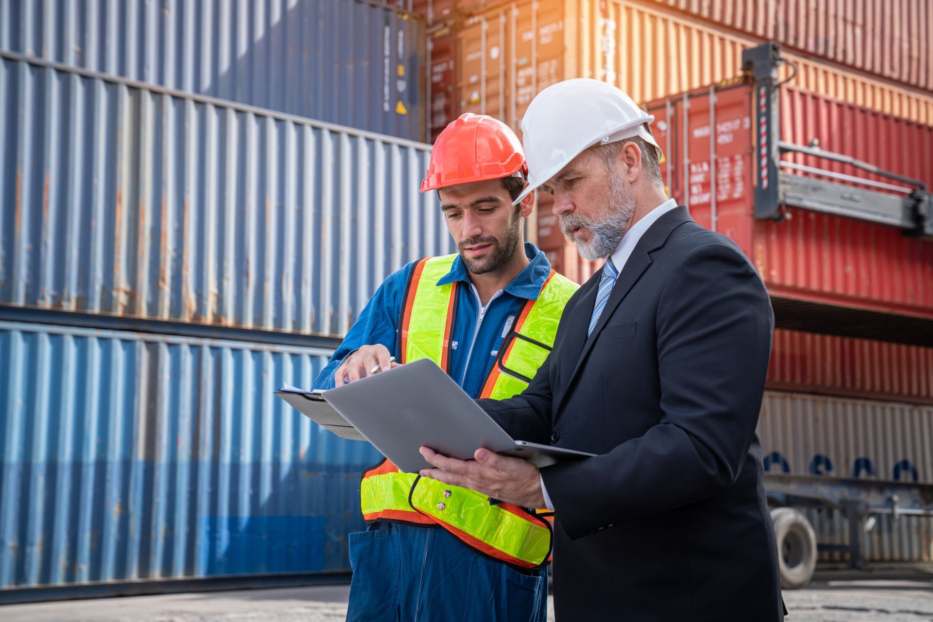 Two workers in hard hats review a clipboard in front of stacked shipping containers.