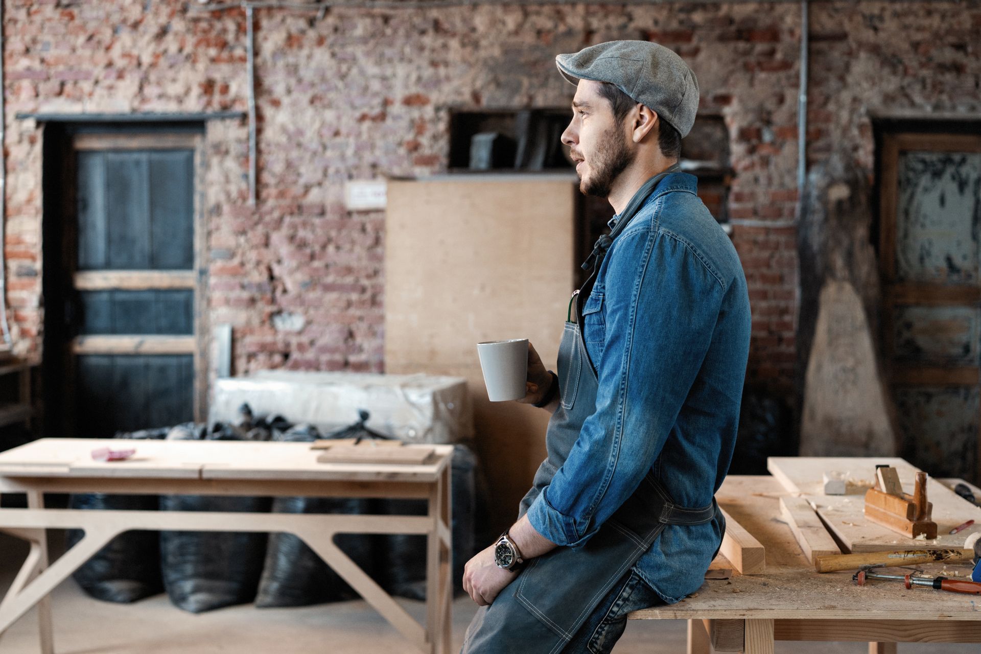 Man in denim shirt and cap sitting in a workshop beside a workbench and tools