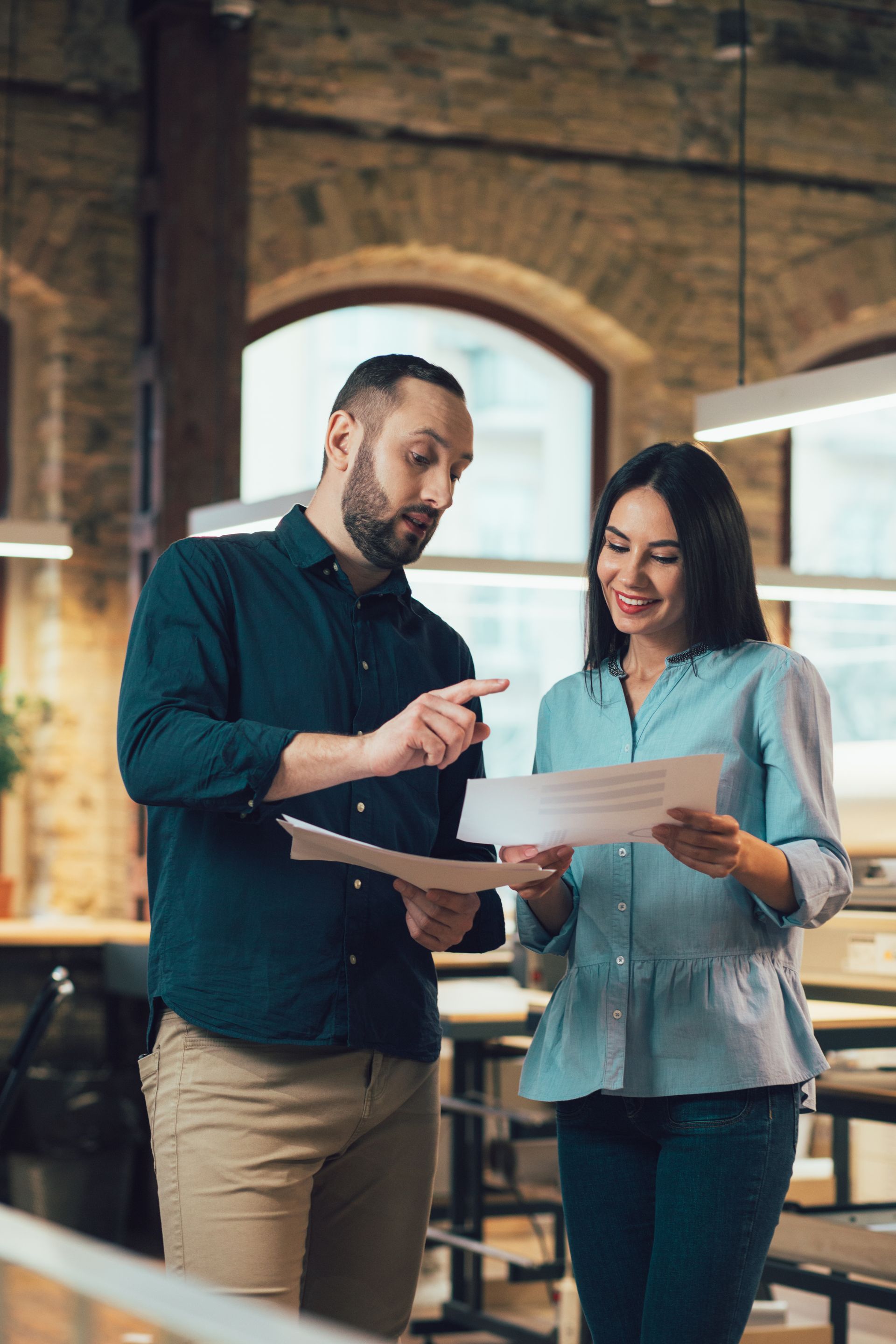 Two colleagues review a document in a brick-walled office, pointing at papers and smiling.