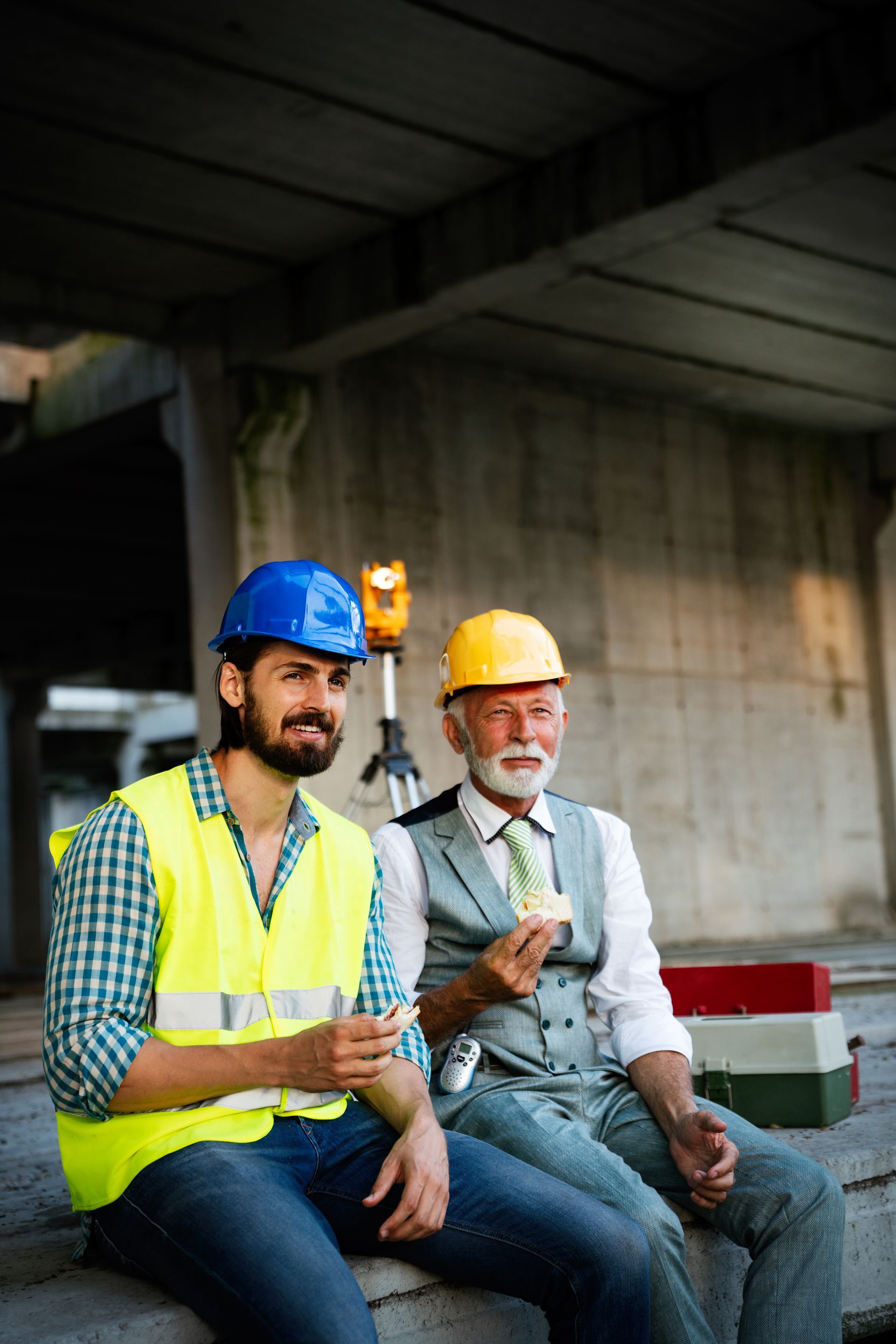 Two construction workers in hard hats and safety vests sit and talk at a building site.
