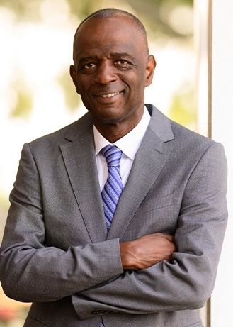 Smiling man in a gray suit with arms crossed, standing indoors against a bright background