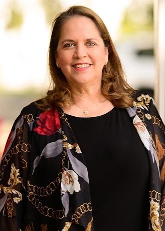 Smiling woman in black top and floral cardigan standing indoors against a blurred background