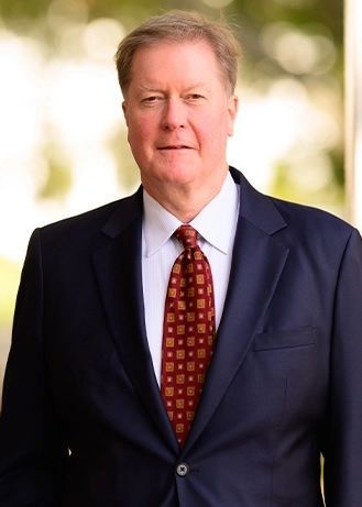 Man in a navy suit and red patterned tie standing against a blurred outdoor background.