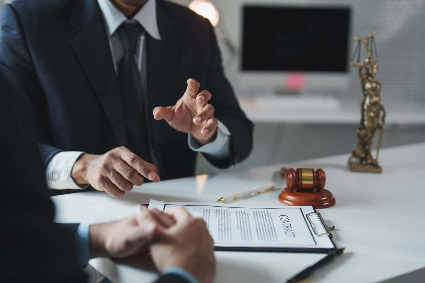 Lawyer in a suit gestures at a desk with contract papers, gavel, and statuette in an office setting