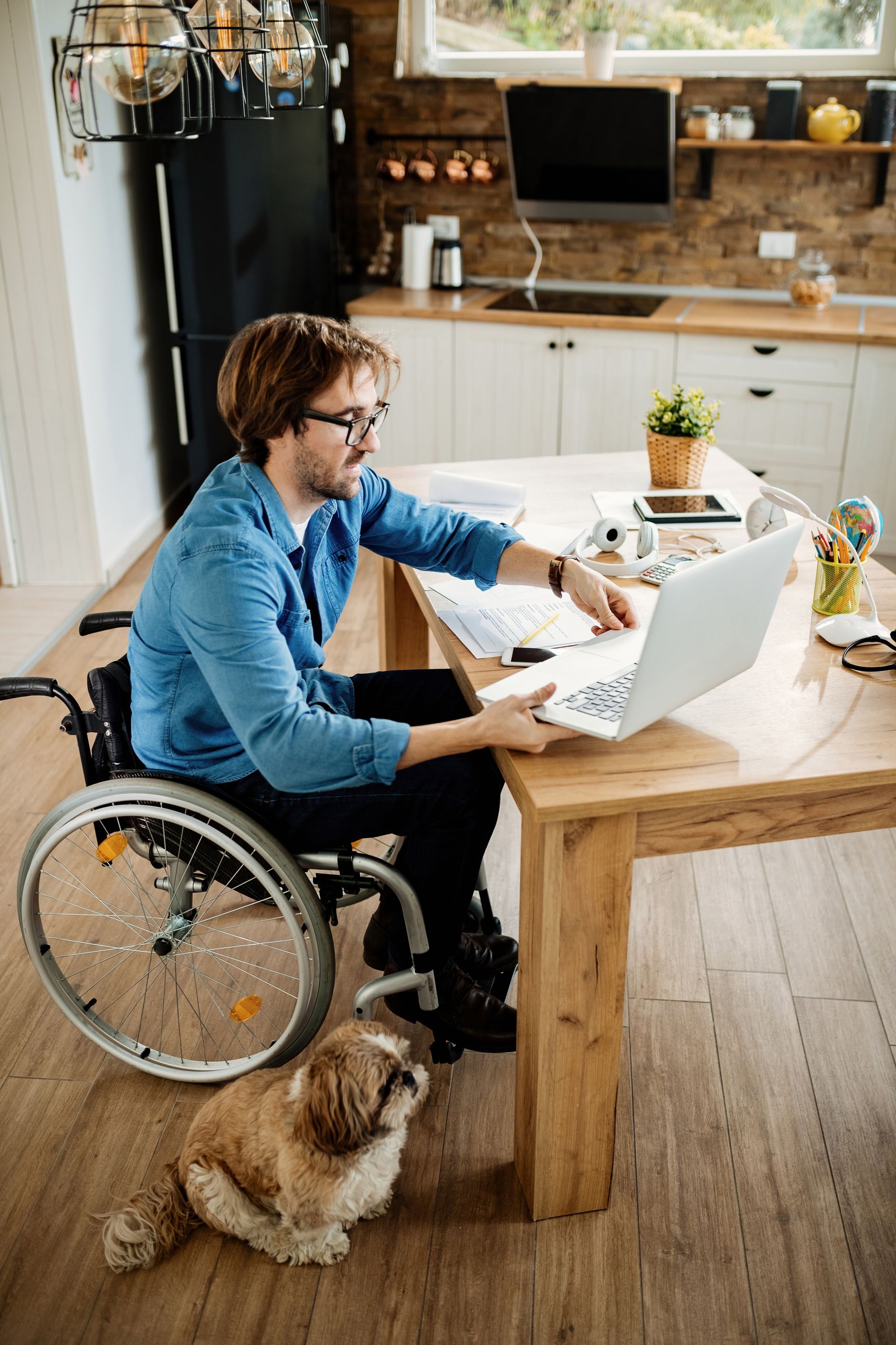 Person in a wheelchair using a laptop at a kitchen table with a dog resting beside them.