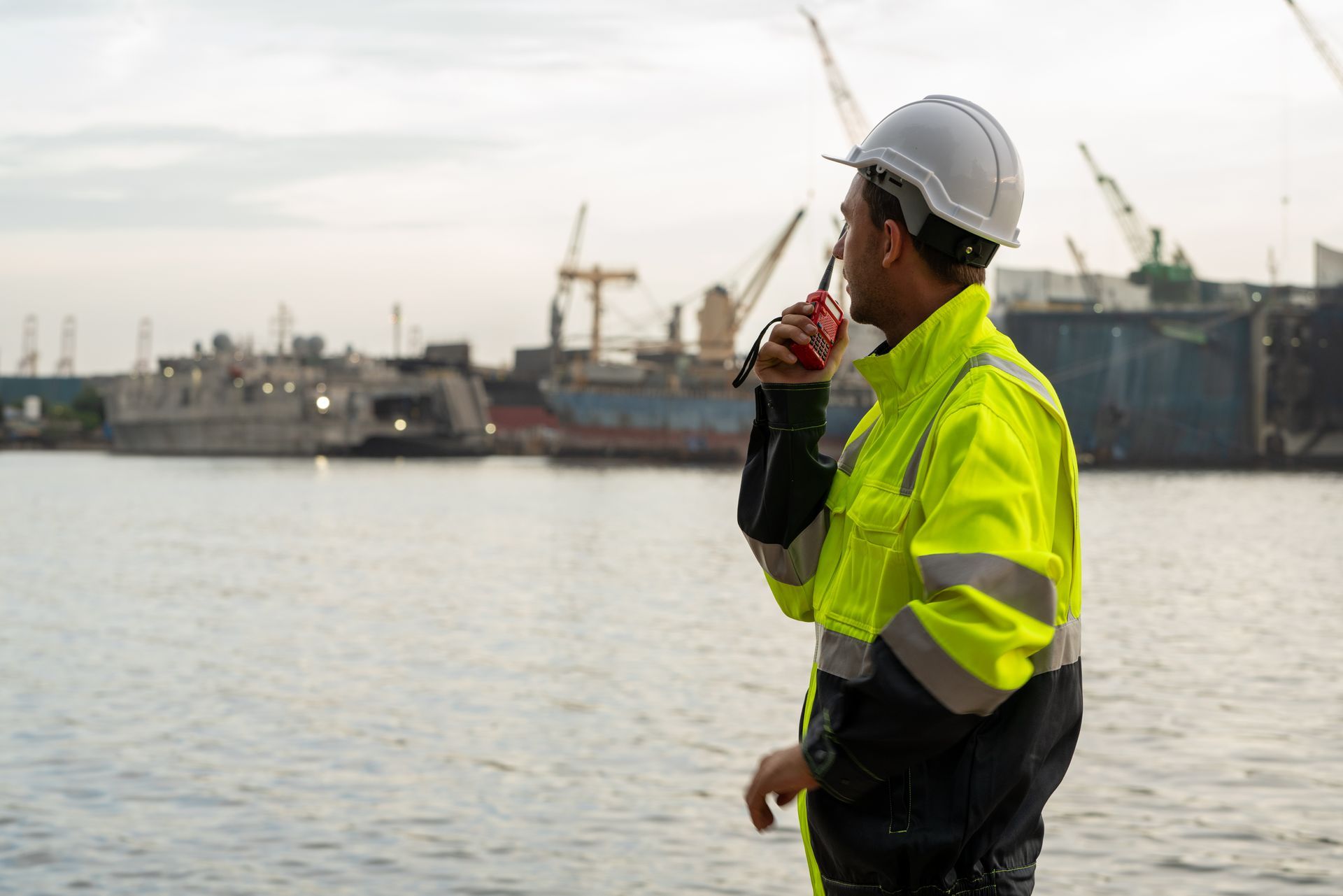 Worker in a hard hat and neon jacket overlooking water at an industrial port with cranes
