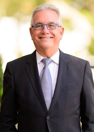 Smiling man in a dark suit and tie standing outdoors against a bright blurred background