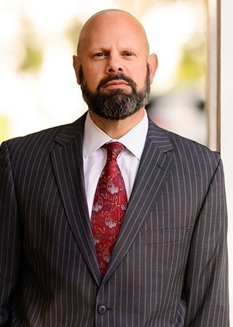 Portrait of a bald man in a gray pinstripe suit with a red tie, standing outdoors.