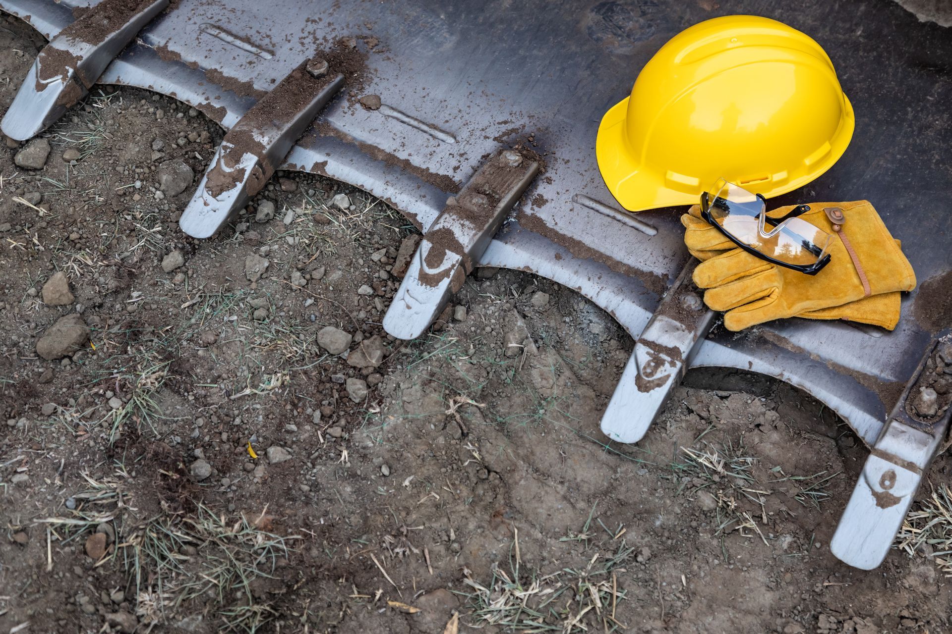 Yellow hard hat and work gloves beside an excavator bucket on dirt ground