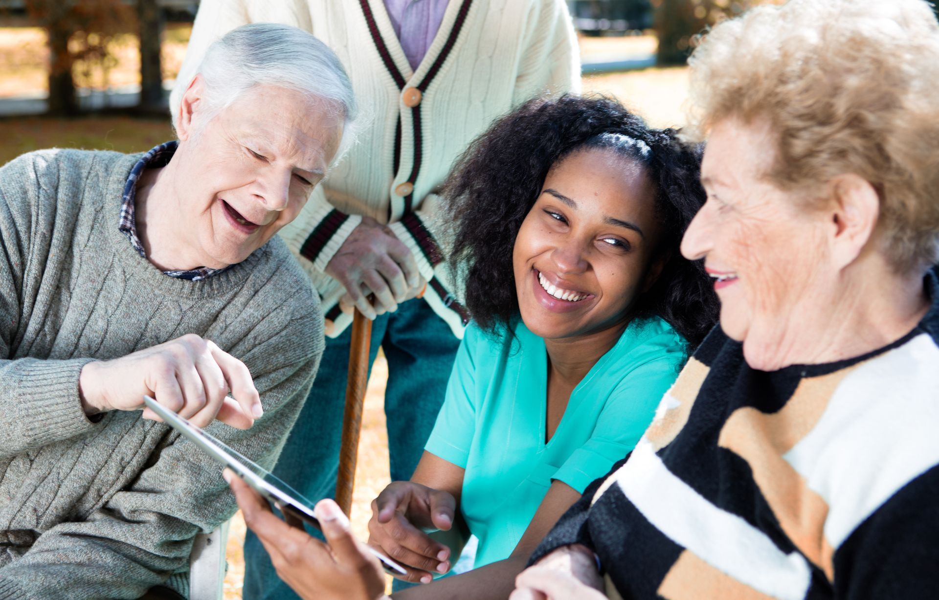 Two seniors and a caregiver looking at a tablet, smiling outdoors.