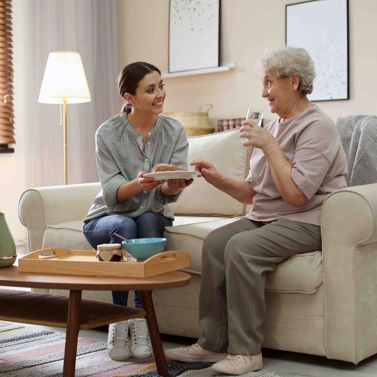 Young woman offering plate to older woman on a sofa, inside a living room.