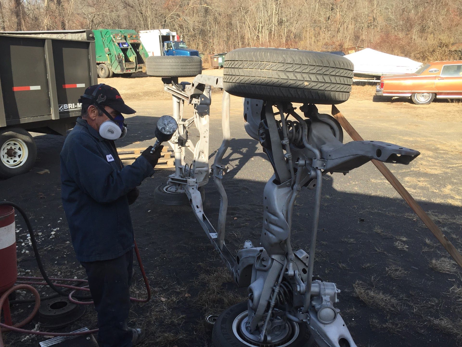 Man sandblasting a car chassis outdoors, wearing a respirator. Near a truck and a classic car. | Rose Brothers Garage