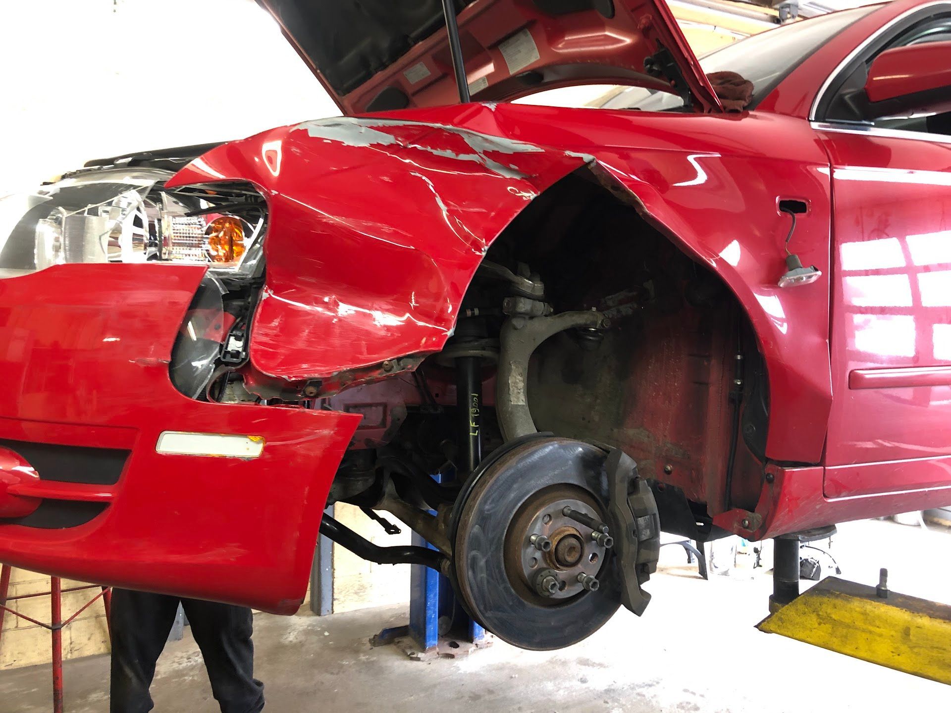 Red car with severe front-end damage, hood open. Front tire removed, revealing suspension components in a repair shop. | Rose Brothers Garage