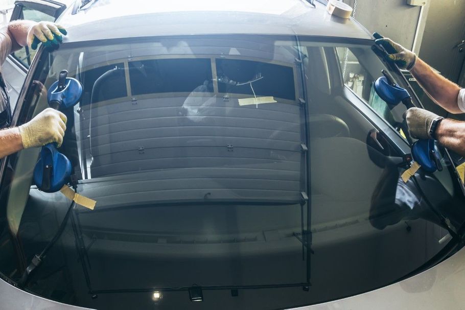 Two workers installing a windshield on a car in a workshop, using blue suction tools.