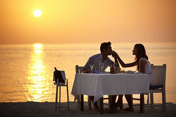 A man and a woman are sitting at a table by the ocean at sunset.