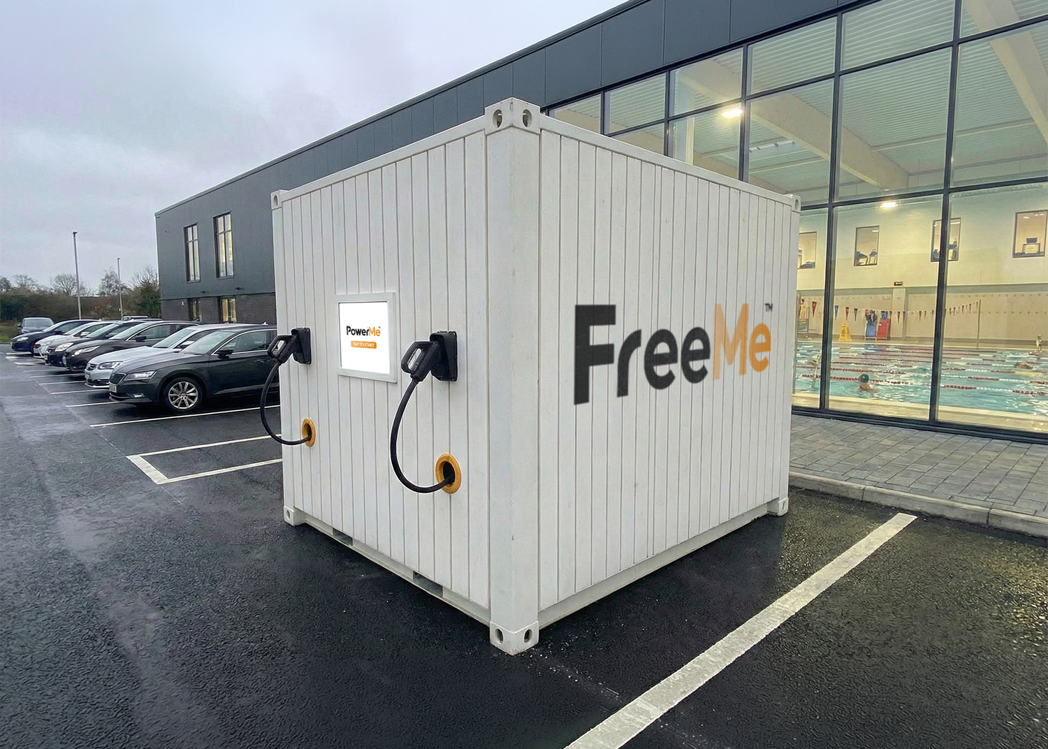 White  ground work free FreeMe EV charging unit in a wet parking lot beside a glass-fronted building