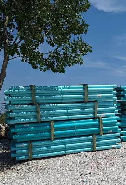 Stacked turquoise pipes beside a tree on a gravel lot under a blue sky